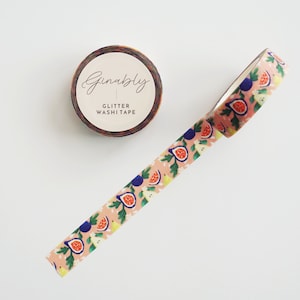May include: A roll of glitter washi tape with a fig pattern and the text "Ginably Glitter Washi Tape." The tape features a repeating design of figs, leaves, and other elements in shades of pink, blue, and green. The roll is brown and white.