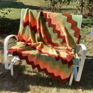 May include: A crocheted blanket featuring a chevron pattern in alternating horizontal stripes of dark brown, sage green, light yellow, and bright orange yarn. The textile is draped over a white metal outdoor rocking chair.