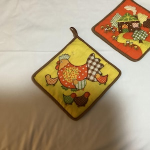 May include: Two square quilted pot holders with brown fabric trim and hanging loops. One yellow pot holder features a central hen with chicks, while the other orange pot holder displays a house, trees, and flowers in a rural landscape design.