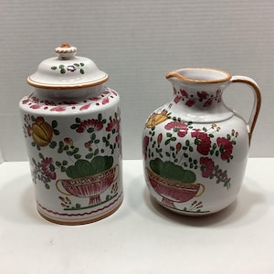 May include: A white ceramic canister with a matching lid and a white ceramic pitcher, both featuring hand-painted floral motifs in red, green, and yellow. The designs include stylized flowers emerging from decorative urns with orange trim.