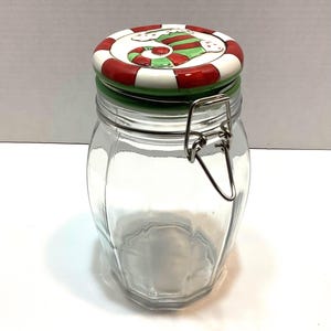 May include: A clear glass storage jar with a metal wire bail closure. The lid is ceramic, featuring a red and white striped border and a central illustration of a green and red striped Christmas stocking with white cuffs on a white background.