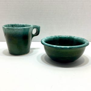 May include: A ceramic mug and a matching bowl featuring a deep forest green glaze with lighter teal and white highlights along the rims. The mug has a wide, circular handle, and both pieces exhibit a smooth, glossy finish with subtle color variations.