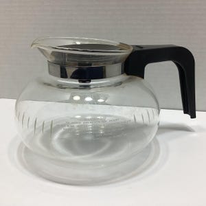 May include: A clear glass coffee carafe with a black plastic handle and a black lid. The glass body features a silver-colored metal band around the top and white printed text that reads WARNING TO AVOID BREAKAGE OR INJURY.