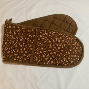 May include: A double oven mitt featuring a brown fabric base with a repeating pattern of small orange flowers and white dots. The mitt has a quilted texture throughout, brown fabric trim along the edges, and a small loop for hanging on one side.