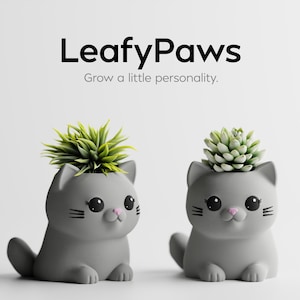 Könnte beinhalten: Zwei graue katzenförmige Pflanzgefäße mit grünen Pflanzen. Die Pflanzgefäße haben schwarze Schnurrhaare, große schwarze Augen und rosa Nasen. Der Text "LeafyPaws" und "Grow a little personality" stehen oben.
