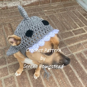 May include: A gray crocheted shark hat for a dog featuring a dorsal fin on top, two black circular eyes, and a row of white triangular teeth along the front edge. The text Crochet Pattern by Serene Homestead is overlaid on the image.