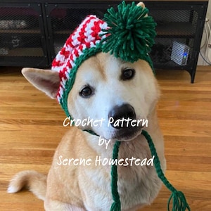 May include: A crochet hat for a dog featuring red and white stripes, a green trim, and a large green yarn pom-pom on top. The hat includes long green braided ties with tassels. The text Crochet Pattern by Serene Homestead is overlaid on the image.