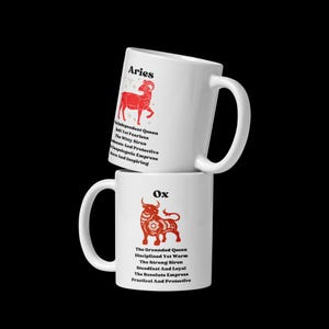 May include: Two white ceramic mugs stacked. The top mug displays "Aries" with a red ram illustration and text. The bottom mug shows "Ox" with a red bull illustration and text. Both mugs have black text. The mugs are set against a black background.