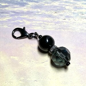 May include: A decorative charm featuring a black metal lobster clasp, a smooth round black bead, a small silver-toned spacer ring set with clear crystals, and a faceted translucent dark gray glass bead at the end of the assembly.