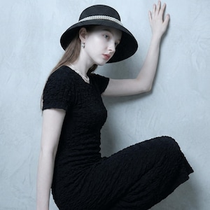 Könnte beinhalten: Ein schwarzer Sonnenhut mit einem perlenartigen Band und breiter Krempe. Der Hut wird mit einem schwarzen strukturierten Kleid getragen. Das Bild zeigt eine Person in einer Pose mit ausgestrecktem Arm.