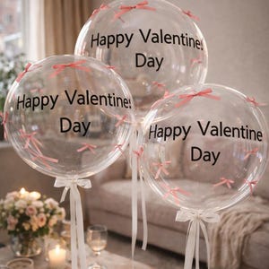 Puede incluir: Tres globos transparentes con la frase "Happy Valentine's Day" en negro. Cada globo está adornado con pequeños lazos rosas y cintas blancas. El fondo borroso sugiere un ambiente festivo y romántico.