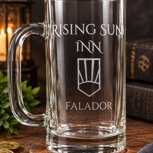 May include: A clear glass beer mug featuring an etched design with the text RISING SUN INN, a shield icon with sun rays, and the word FALADOR below. The mug has a sturdy handle and a thick base, designed for holding beverages in a classic style.