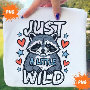 Puede incluir: Una camiseta blanca con una ilustración gráfica de un mapache gris y negro. El texto dice JUST A LITTLE WILD en letras negritas. El diseño está rodeado de pequeños corazones rojos y estrellas azul claro, creando un visual detallado y divertido.
