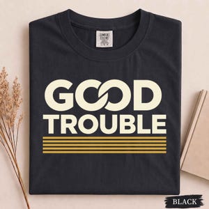 May include: A black short-sleeved t-shirt featuring the text "GOOD TROUBLE" in large, cream-colored capital letters. Below the text are five horizontal gold lines. The shirt has a Comfort Colors brand label at the inner neckline.