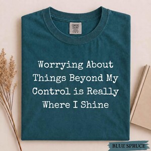 May include: A dark teal crew neck t-shirt with the white text "Worrying About Things Beyond My Control is Really Where I Shine" printed on the front. The shirt features a Comfort Colors brand label on the inside of the collar.