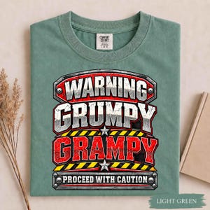 May include: A sage green short-sleeve t-shirt featuring a graphic design with the text WARNING GRUMPY GRAMPY PROCEED WITH CAUTION. The design includes metallic-style lettering, red and black hazard stripes, and two silver stars on a dark background.