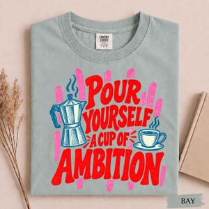 K&ouml;nnte beinhalten: Ein hellgraues T-Shirt mit einem Grafikdruck einer Mokkakanne und einer Kaffeetasse sowie dem Text "Pour Yourself A Cup Of Ambition" in fetten roten Buchstaben. Das Design enth&auml;lt rosa vertikale Pinselstriche und ein Markenetikett.