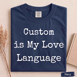 May include: A navy blue crew neck t-shirt featuring the white text "Custom is My Love Language" printed on the front. The shirt includes a Comfort Colors brand label at the inner neckline. The design uses a bold, distressed-style white font.