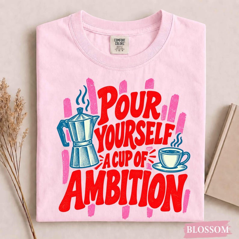 K&ouml;nnte beinhalten: Ein hellrosa T-Shirt mit einem Grafikdruck einer blauen Mokkakanne und einer Kaffeetasse. Der Text "POUR YOURSELF A CUP OF AMBITION" ist in fetten roten Buchstaben gedruckt. Das Design enth&auml;lt vertikale rosa Pinselstrich-Akzente hinter dem Text.