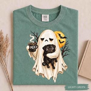 May include: A sage green t-shirt featuring a graphic of a white ghost holding a black mug with the text MEH and embracing a black pug. The design includes a yellow moon and black silhouettes of bats and bare tree branches against the fabric.