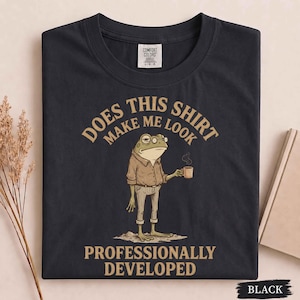 May include: A black short-sleeve t-shirt featuring a graphic of a frog wearing a button-down shirt and glasses while holding a coffee mug. The text reads: DOES THIS SHIRT MAKE ME LOOK PROFESSIONALLY DEVELOPED. The shirt has a Comfort Colors label.