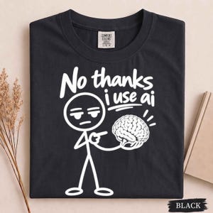 May include: A black t-shirt featuring a white graphic of a stick figure pointing at a brain illustration. The text above the figure reads "No thanks i use ai" in a bold, white, hand-drawn style font. The shirt has a Comfort Colors brand label.