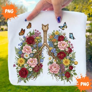Puede incluir: Sudadera blanca con un diseño floral de pulmones. El diseño presenta flores rojas, rosas, amarillas y azules, ramas marrones y tres mariposas. La sudadera es sostenida por una persona.