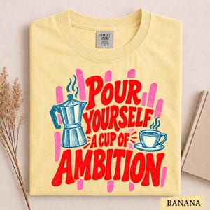 K&ouml;nnte beinhalten: Ein hellgelbes Kurzarm-T-Shirt mit einem Grafikdruck einer Mokkakanne und einer Kaffeetasse mit Dampf. Der Text "POUR YOURSELF A CUP OF AMBITION" ist in fetten roten Buchstaben gedruckt, akzentuiert durch rosa vertikale Pinselstriche im Hintergrund.