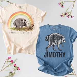 Pode incluir: Uma t-shirt creme com um guaxinim, um arco-íris e o texto different is beautiful, ao lado de uma t-shirt azul com um guaxinim e o texto JIMOTHY. Ambas as t-shirts têm mangas curtas e gola redonda com estampas gráficas na parte frontal.