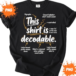 Op de afbeelding: Een zwart T-shirt met korte mouwen en witte en gouden tekst die This shirt is decodable leest. Het ontwerp bevat diverse taalkundige annotaties met pijlen die naar specifieke woorden wijzen, waarbij concepten zoals digrafen, gesloten lettergrepen en achtervoegsels worden uitgelegd.