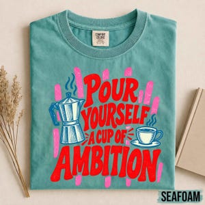 K&ouml;nnte beinhalten: Ein blaugr&uuml;nes T-Shirt mit Rundhalsausschnitt und einem Grafikdruck einer Mokkakanne sowie einer Kaffeetasse mit dem Text "Pour yourself a cup of ambition" in roter Schrift. Das Design enth&auml;lt rosa vertikale Pinselstriche hinter dem Text.