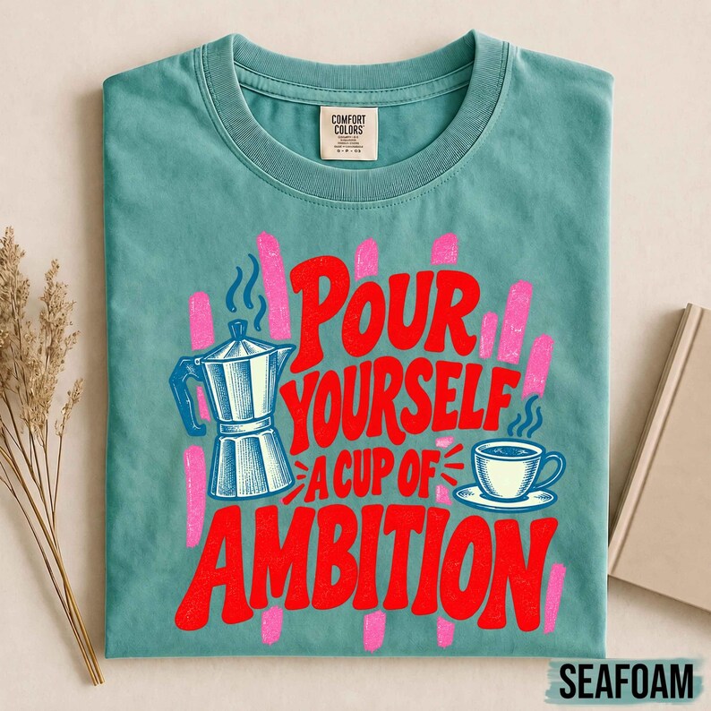 K&ouml;nnte beinhalten: Ein blaugr&uuml;nes T-Shirt mit Rundhalsausschnitt und einem Grafikdruck einer Mokkakanne sowie einer Kaffeetasse mit dem Text "Pour yourself a cup of ambition" in roter Schrift. Das Design enth&auml;lt rosa vertikale Pinselstriche hinter dem Text.