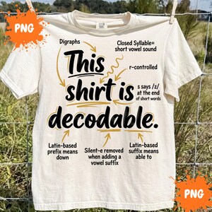 Op de afbeelding: Een crèmekleurig t-shirt met korte mouwen en zwarte en gouden tekst die luidt: This shirt is decodable. Het ontwerp bevat diverse taalkundige annotaties en pijlen die naar specifieke woorden wijzen, met uitleg over fonetische regels zoals digrafen, r-gecontroleerde klinkers en Latijnse voor- en achtervoegsels.