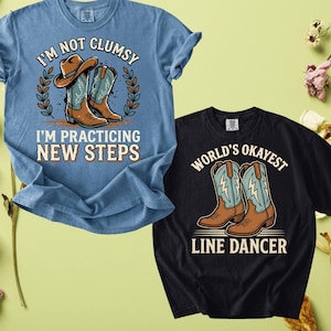 May include: A blue t-shirt with a graphic of brown cowboy boots and the text I'M NOT CLUMSY I'M PRACTICING NEW STEPS, and a black t-shirt with a graphic of brown cowboy boots and the text WORLD'S OKAYEST LINE DANCER.