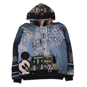 Op de afbeelding: Een sweatshirt met lange mouwen en capuchon met een geweven tapijtontwerp. De voorkant toont een groot kasteel, vuurwerk en een Mickey Mouse-figuur. De capuchon en het onderste gedeelte tonen kleine figuren tegen een zwarte achtergrond.