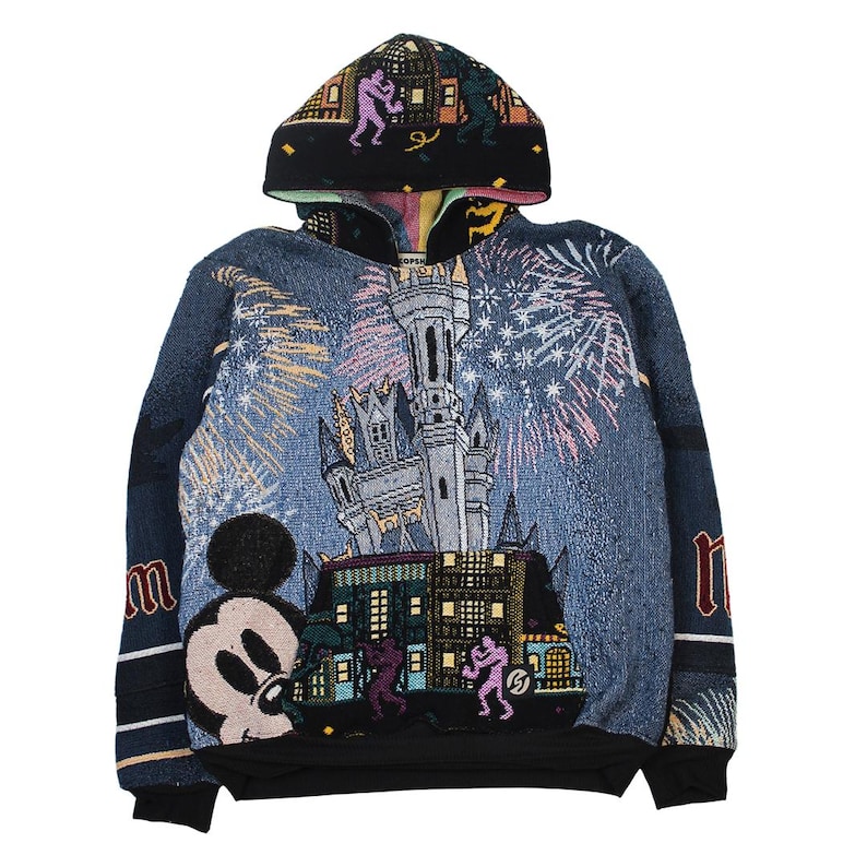 Op de afbeelding: Een sweatshirt met lange mouwen en capuchon met een geweven tapijtontwerp. De voorkant toont een groot kasteel, vuurwerk en een Mickey Mouse-figuur. De capuchon en het onderste gedeelte tonen kleine figuren tegen een zwarte achtergrond.