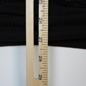Op de afbeelding: Een zwarte geribbelde stoffen rand van een kledingstuk naast een houten liniaal. De liniaal toont metingen in centimeters, waarbij de zoom van de stof bij de 63,5 cm markering ligt en voorbij de 68,5 cm markering reikt, wat een lengte van ruim 5 cm aangeeft.