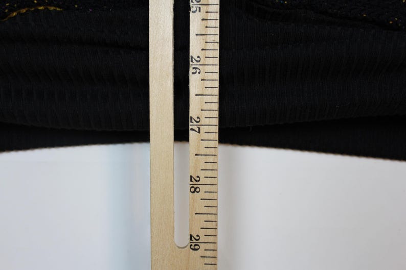 Op de afbeelding: Een zwarte geribbelde stoffen rand van een kledingstuk naast een houten liniaal. De liniaal toont metingen in centimeters, waarbij de zoom van de stof bij de 63,5 cm markering ligt en voorbij de 68,5 cm markering reikt, wat een lengte van ruim 5 cm aangeeft.