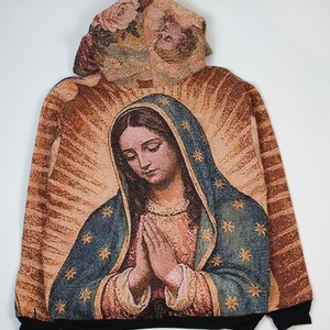 May include: A long-sleeve hoodie featuring a woven tapestry-style graphic of the Virgin Mary with hands clasped in prayer. The design includes a blue cloak with gold stars, a tan background with radiating light rays, and a floral pattern on the hood.