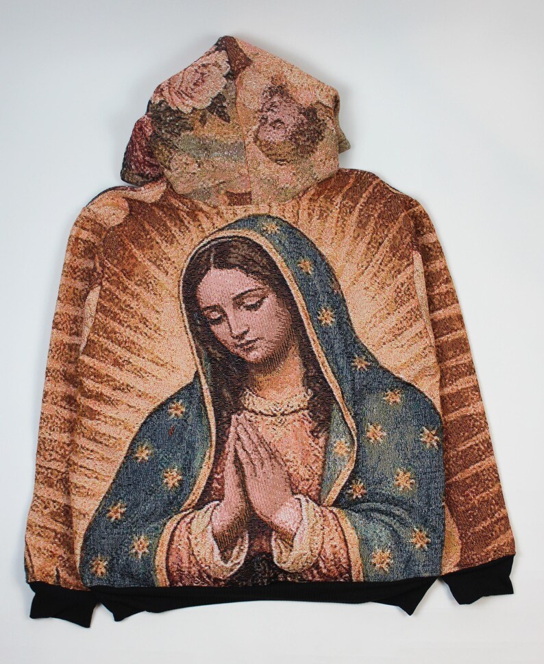 May include: A long-sleeve hoodie featuring a woven tapestry-style graphic of the Virgin Mary with hands clasped in prayer. The design includes a blue cloak with gold stars, a tan background with radiating light rays, and a floral pattern on the hood.