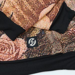May include: A long-sleeve sweatshirt featuring a woven tapestry-style floral pattern in shades of copper, brown, and muted pink. The garment includes solid black ribbed cuffs and a black ribbed hem, with a small square black patch displaying a white logo.