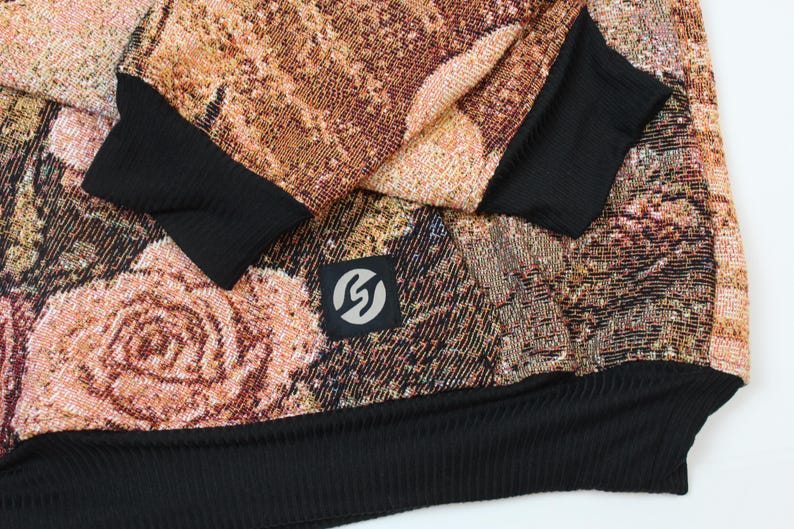 May include: A long-sleeve sweatshirt featuring a woven tapestry-style floral pattern in shades of copper, brown, and muted pink. The garment includes solid black ribbed cuffs and a black ribbed hem, with a small square black patch displaying a white logo.
