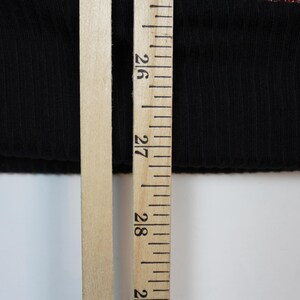 May include: A black ribbed fabric garment edge positioned next to a wooden ruler. The ruler shows a measurement of approximately 27 inches, providing a clear scale for the length of the black textile material visible in the frame.