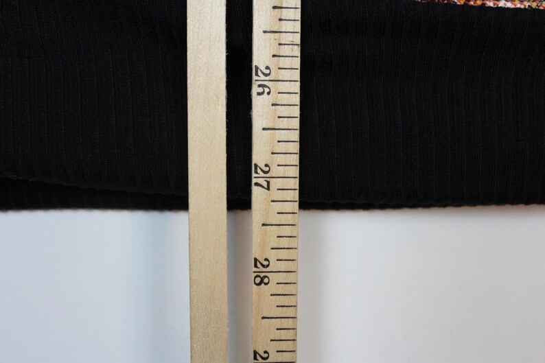 May include: A black ribbed fabric garment edge positioned next to a wooden ruler. The ruler shows a measurement of approximately 27 inches, providing a clear scale for the length of the black textile material visible in the frame.