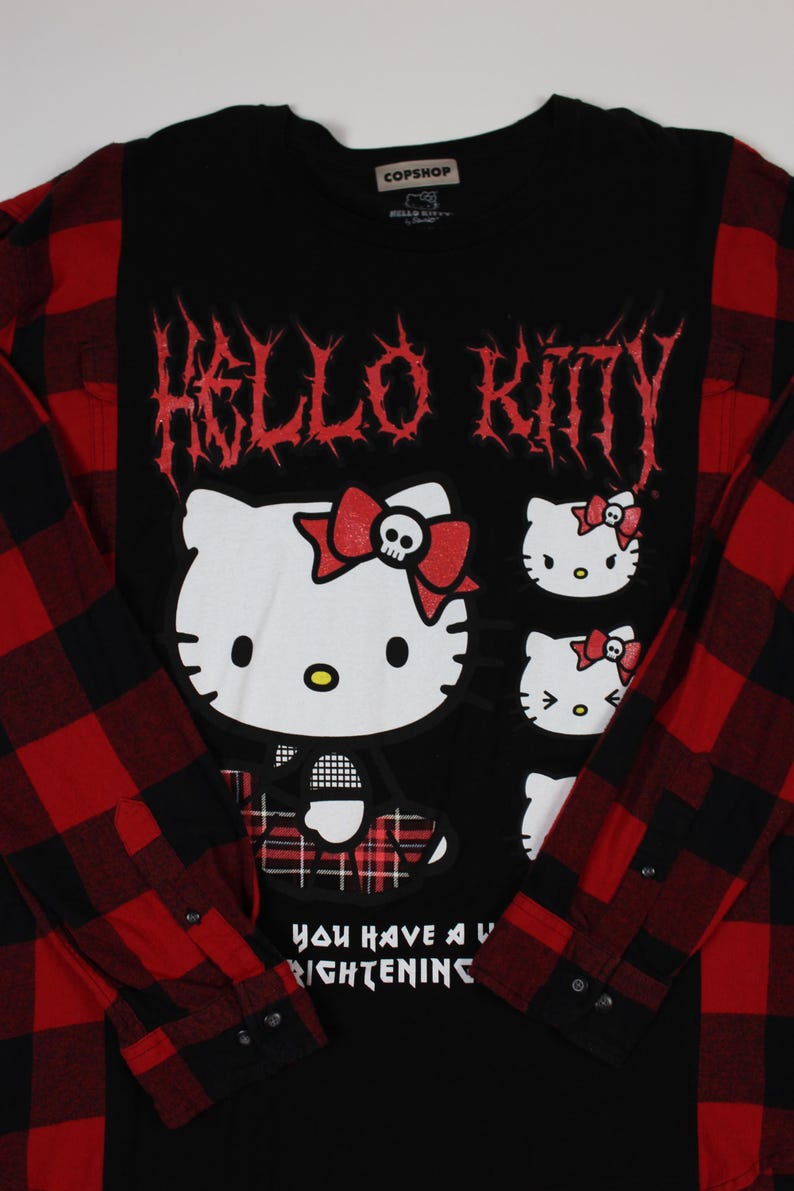 May include: Black long-sleeved shirt with a red and black plaid pattern on the sleeves. The shirt features a graphic of Hello Kitty with a skull bow and the text "Hello Kitty" in red. The shirt also has the text "You have a frightening" on the bottom.