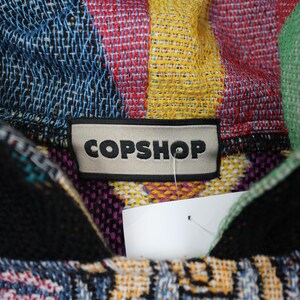 Op de afbeelding: Een veelkleurige geweven hoodie met een opvallend rechthoekig label met de tekst COPSHOP in zwarte hoofdletters. De stof vertoont een levendig, ingewikkeld patroon met tinten blauw, geel, rood, groen en zwart door het hele weefsel heen.