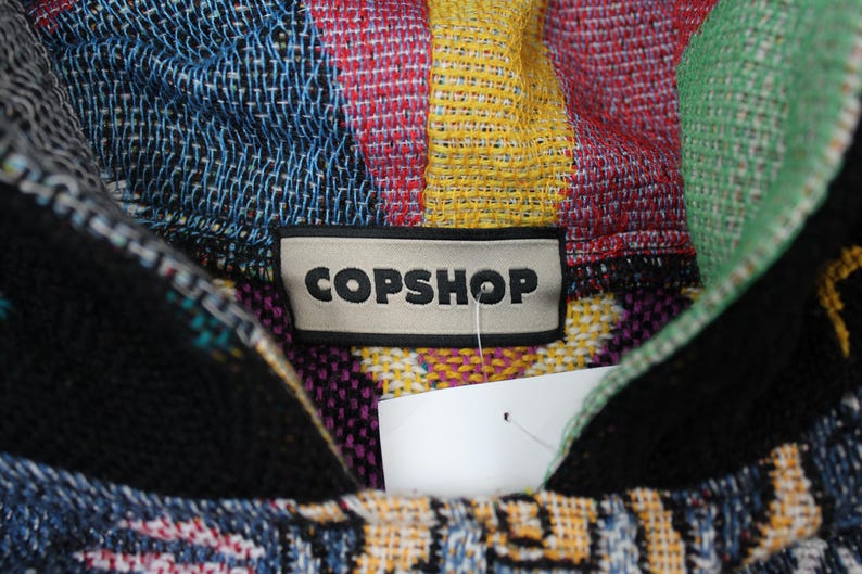 Op de afbeelding: Een veelkleurige geweven hoodie met een opvallend rechthoekig label met de tekst COPSHOP in zwarte hoofdletters. De stof vertoont een levendig, ingewikkeld patroon met tinten blauw, geel, rood, groen en zwart door het hele weefsel heen.