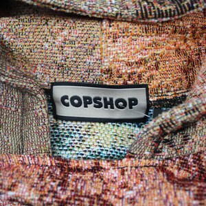 May include: A woven hoodie featuring a multi-colored tapestry pattern in shades of orange, brown, and cream. A rectangular black and white label is stitched at the neckline, displaying the text COPSHOP in bold, black, capital letters.