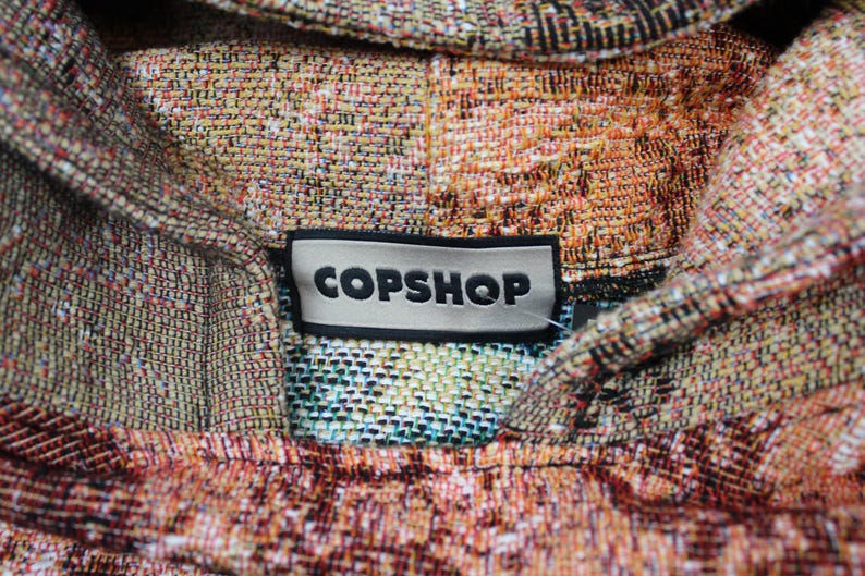 May include: A woven hoodie featuring a multi-colored tapestry pattern in shades of orange, brown, and cream. A rectangular black and white label is stitched at the neckline, displaying the text COPSHOP in bold, black, capital letters.