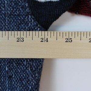 Op de afbeelding: Een blauw en zwart gebreid kledingstuk met een geweven textuurpatroon. Een houten liniaal is horizontaal over de stof geplaatst, waarbij de maatstreepjes van 55 tot 69 centimeter duidelijk zichtbaar zijn en de breedte van het item aangeven.